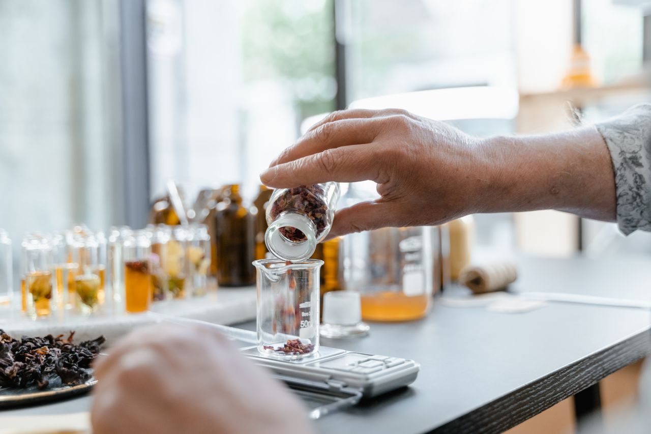 Home Detailed view of hands adding dried botanicals to a glass beaker in a lab setting.
