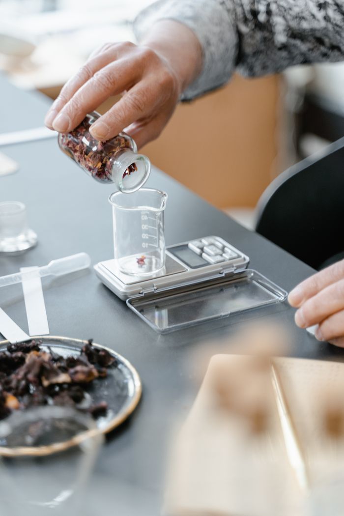 A close-up of hands measuring dried flowers into a beaker on a digital scale, demonstrating laboratory aromatherapy preparation.